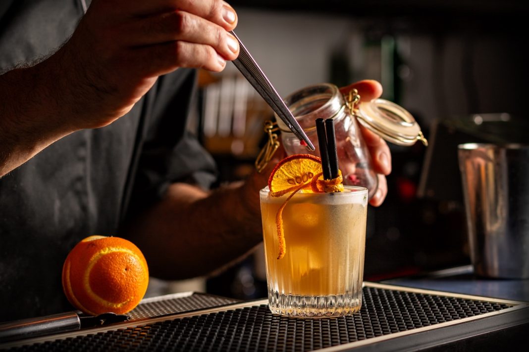 Preparing Smoky Spiced Orange Cocktail decorated with Dehydrated Orange Slices anf fresh orange peel. Bar or pub interior. cocktail