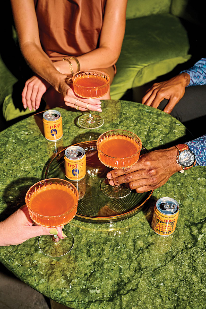 cocktails Three glasses and three cans on a green circular table; the yellow cans sport the title 