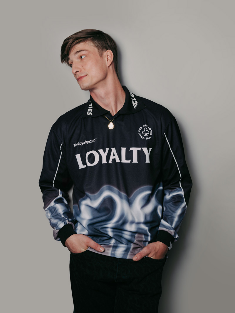Male model wearing dark The Loyalty Club branded racing jersey, wearing black pants. He is leaning on the wall behind him and looking to his right with his hands in his pocket.