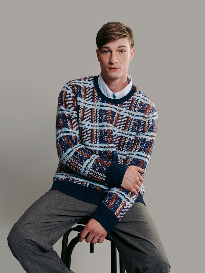 Male model sitting in stool, looking into camera. He is wearing a navy, red, white, and light blue plaid pattern pullover sweater and dark gray trousers.