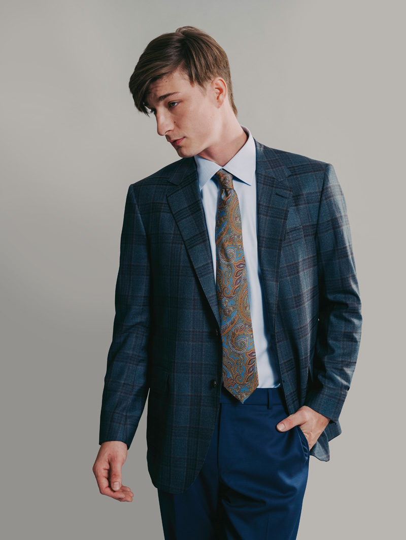Male model is standing with left hand in pants pocket, looking to his right. He is wearing a medium gray blue plaid suit jacket, dark blue pants, and a paisley tie.