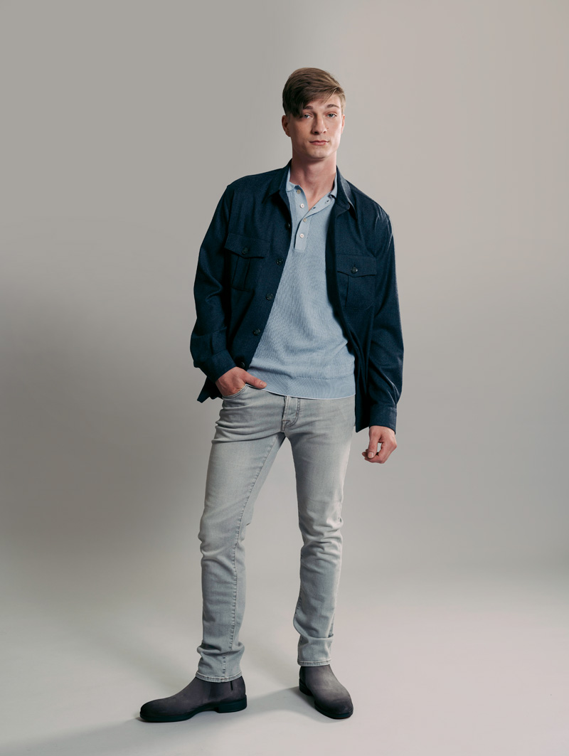 Male model staring into camera with right hand in pocket. He is wearing a dark blue work shirt and alight blue polo underneath. He is also sporting light wash jeans and low steel gray boots.