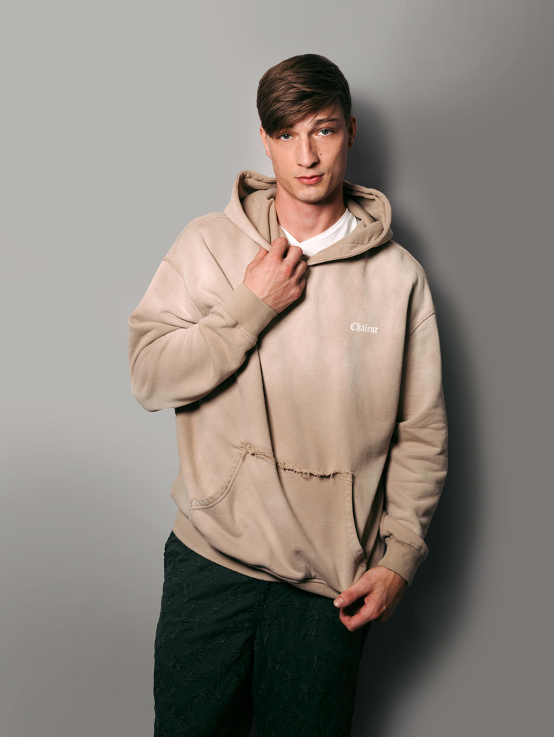 Male model is against the wall, staring into camera. He is wearing a slightly distressed beige hoodie (Chaleur branded on his left pocket area) and dark green embroidered pants.