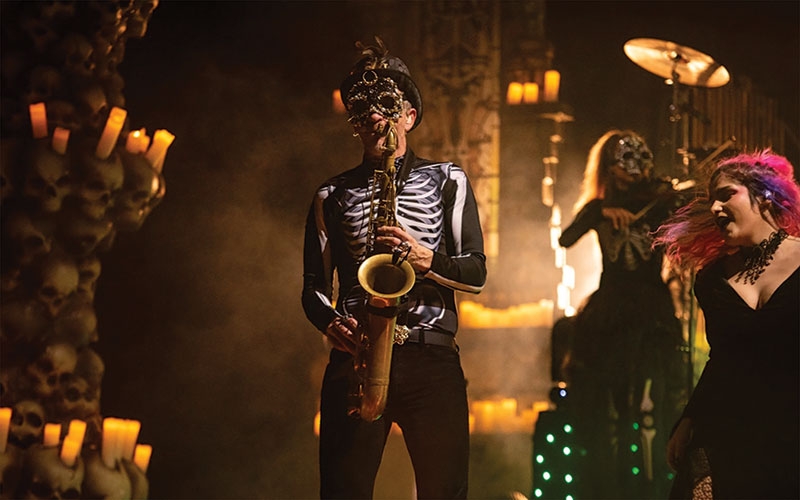 Events Man on stage, playing the saxophone. He is wearing a skull mask and a skeletal ribcage shirt with black pants. To his left behind him are other performers. Another also in a similar outfit playing the violin.
