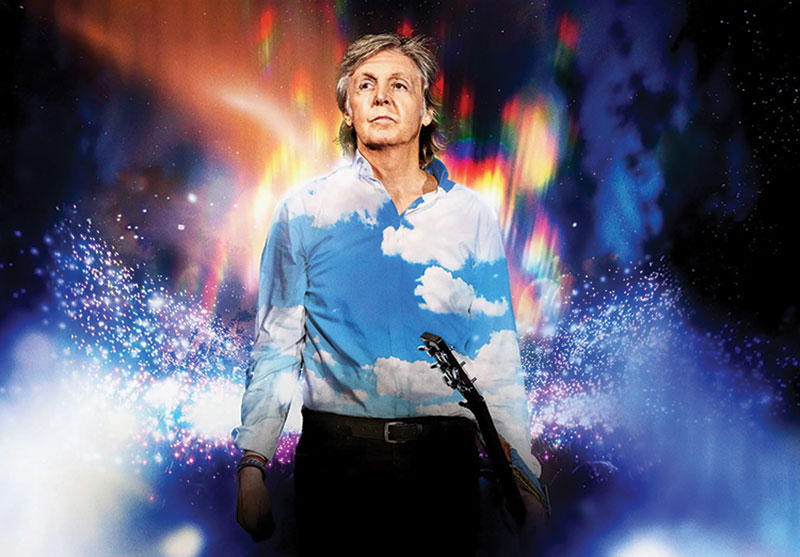 Paul McCartney on stage, wearing a blue shirt with clouds all over. He is holding a guitar, and there are concert lights flashing behind him.