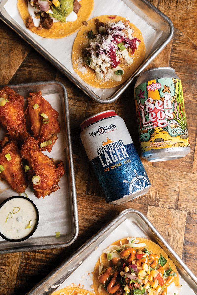 Breweries Two Port of New Orleans Brewing Co. cans on a table, alongside trays of tacos and wings. The featured cans are their Riverfront Lager and their Sea Legs THC-infused Strawberry Lemonade