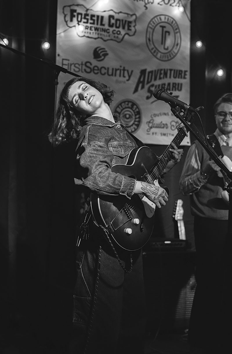 Maddy Kirgo shot in black and white. She's playing her guitar on stage while throwing her head back to the music.