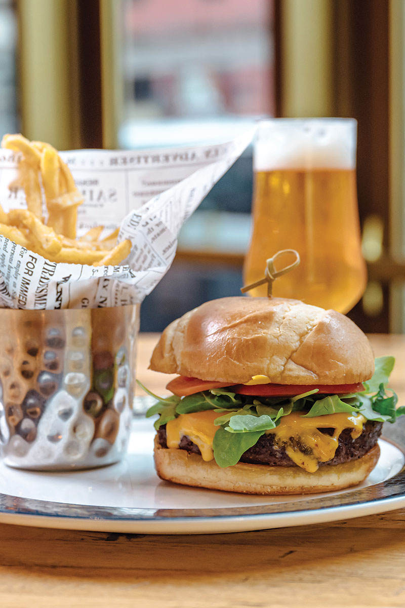 Cheeseburger on plate with toothpick through center. Fries in a metal basket to the left the burger. Both on plate.