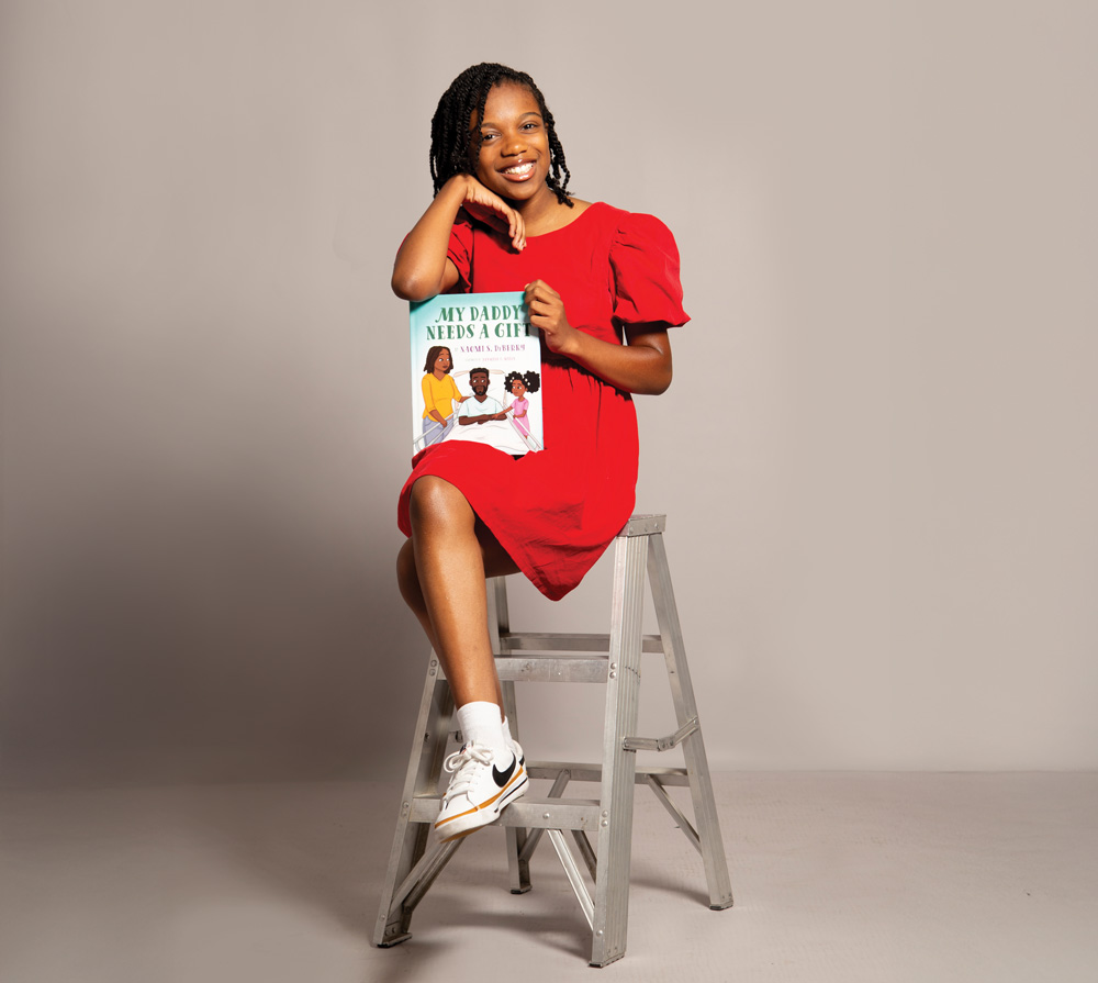 Portrait of Naomi DeBerry. She is a young Black girl. In the photo she is wearing a red dress with white sneakers and holding her book "My Daddy Needs a Gift" with her left hand, and leaning on the book with her right arm. In the photo she is also sitting on a small ladder.