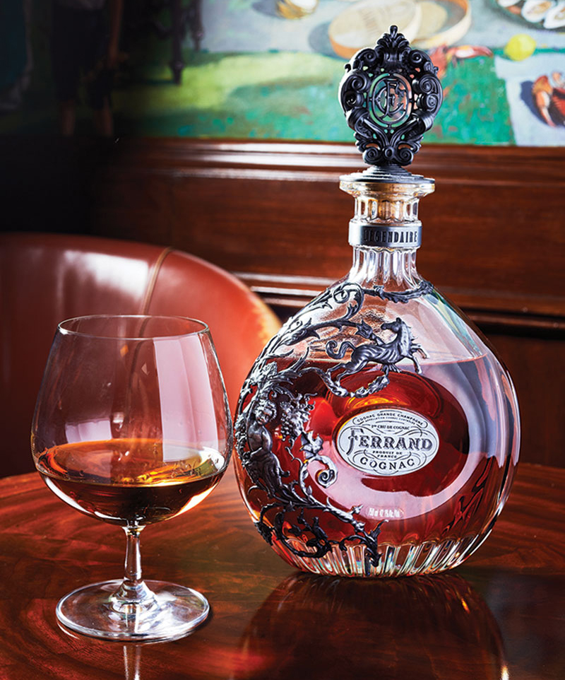 On top of a wooden table: Cognac bottle next to a glass with some Cognac inside the glass.