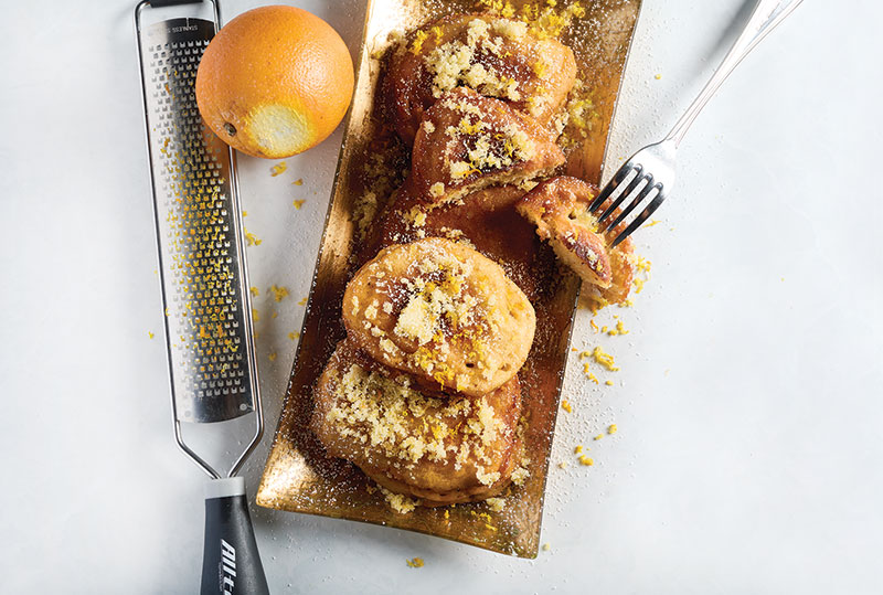 Zeppole Photo of Madeira Flan plated on a long rectangular plate. There is a fork puncturing a piece of the flan to the right of the plate, and a grater with a slightly grated orange to the left of the plate