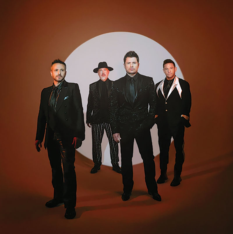 98 Degrees (four member boy band) promotional image –– all 4 men are wearing suits and standing in front of a brown background, with a light circle in the middle