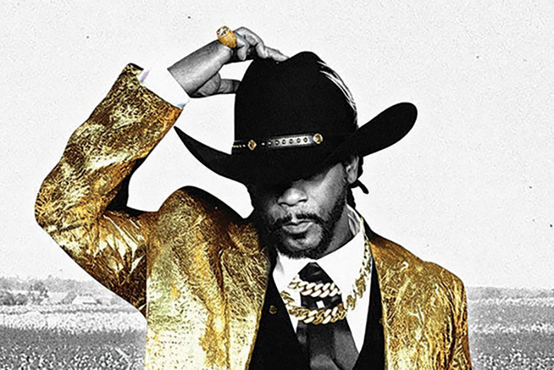 Black and white photo of Katt Williams, save for his gold chains and his gold suit top. He is wearing a black cowboy hat and slightly tipping it down