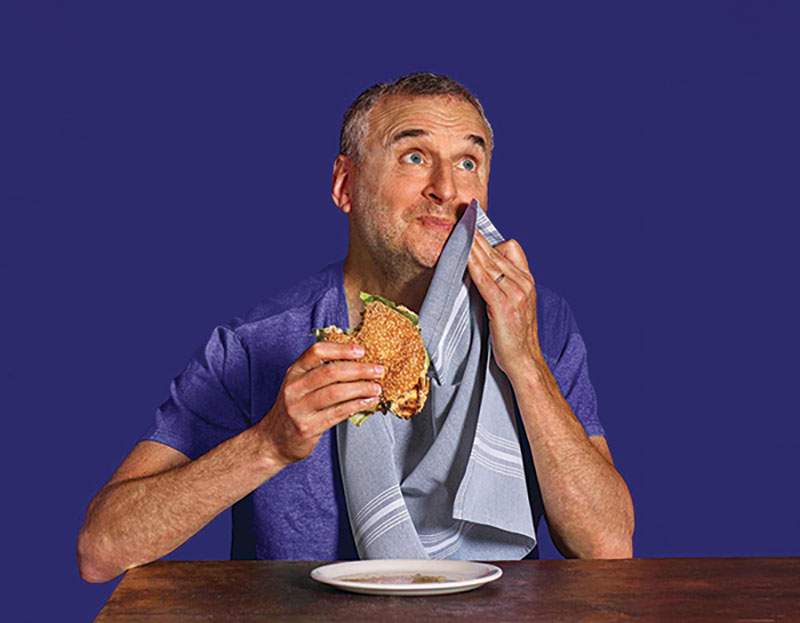 Headshot of Phil, eating a hambuger with his right hand, patting his mouth with a napkin in left hand, looking upwards, left