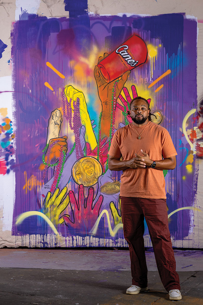 Brandan “BMike” Odums standing in front of his collaboration piece with Cane's. The artwork features a predominantly purple background populated with multiple hands of various colors (i.e. orange-brown, yellow, pink, and a dog paw) The top most hand is holding a Cane's branded cup.